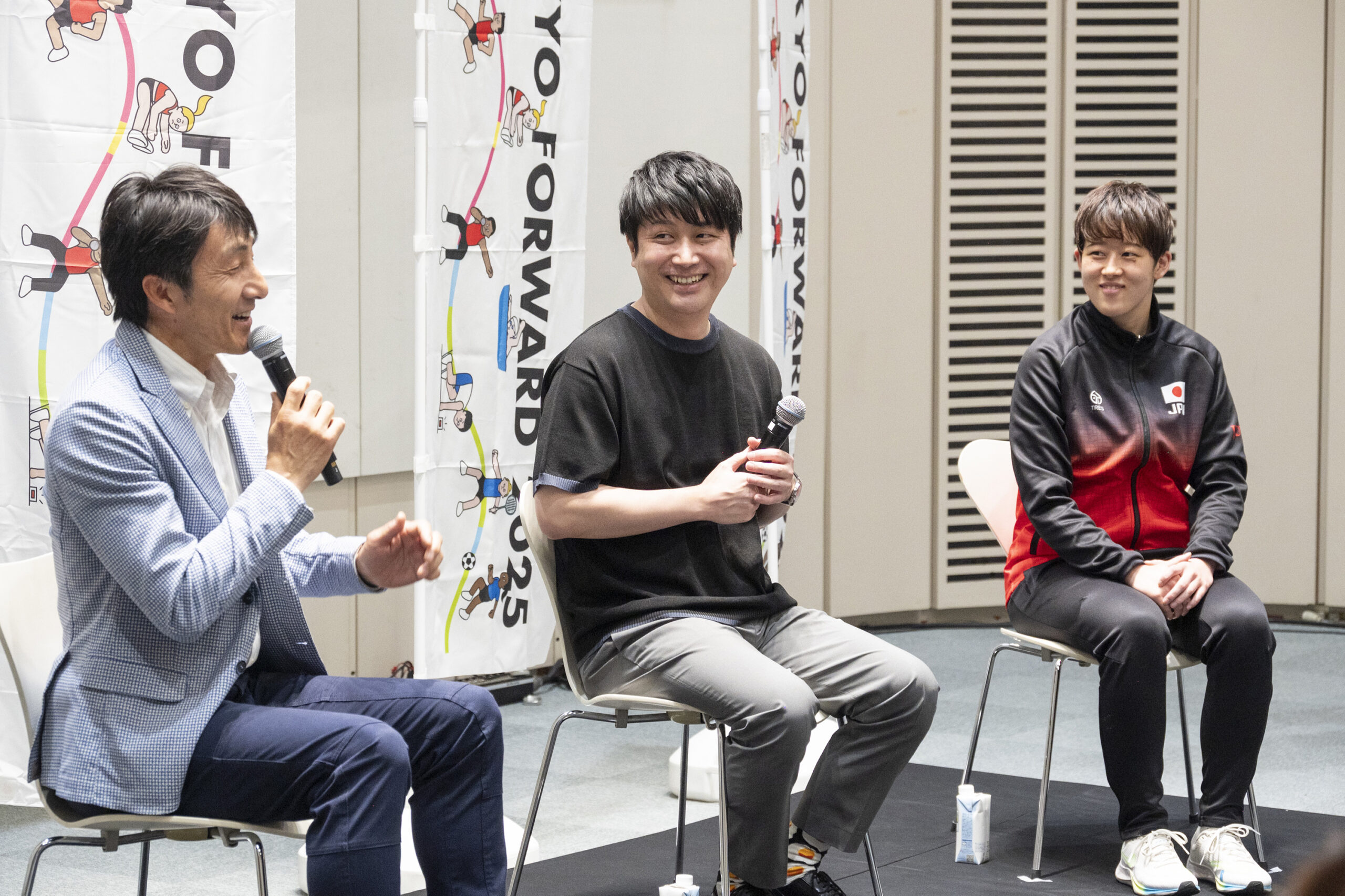 マイクを持って話をしている朝原さんと微笑んで見つめている五十嵐さんと中田選手の写真