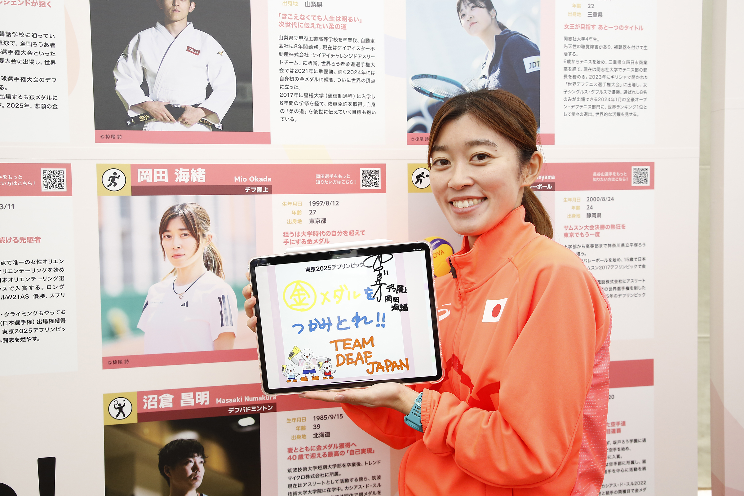 自分のパネルの前でメッセージが書かれたタブレットを持っている岡田選手の写真