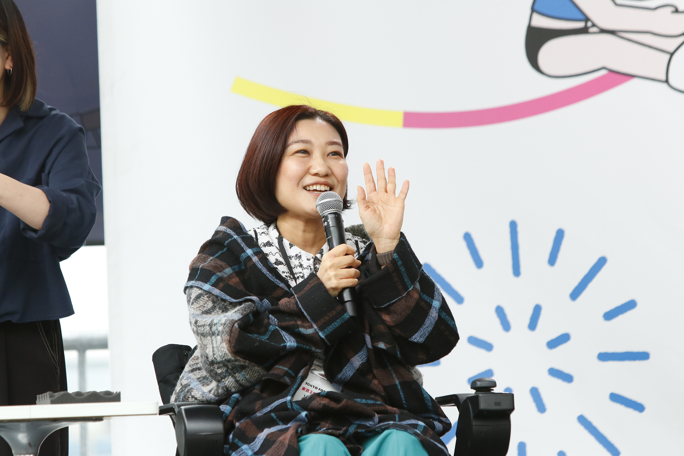 車椅子に乗り左手を顔の横まであげている来栖さんの写真