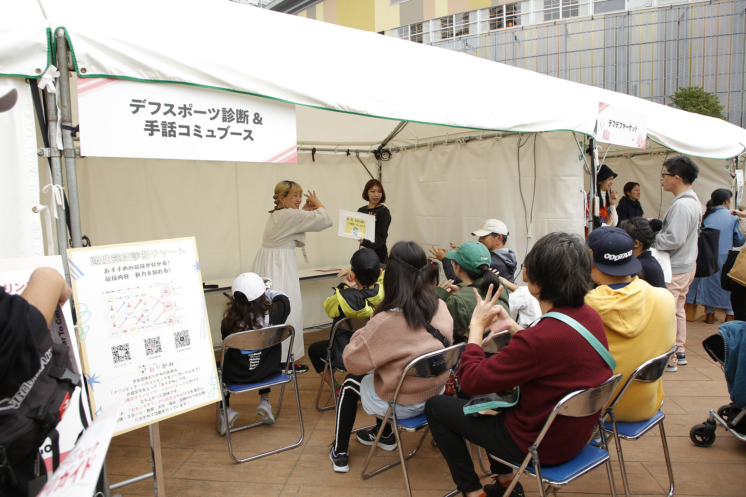 屋外のブース内で女性二人がデフリンピックの手話を参加者に教えている写真